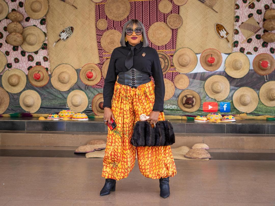 Orange Ankara harem trousers styled with a corset belt against a woven-hat backdrop